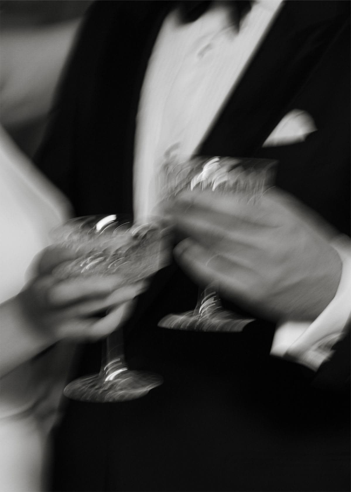 Duas mãos erguendo taças de cristal, smoking e vestido em movimento